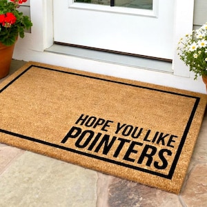 May include: A rectangular doormat made of natural coir fiber. The mat has a black border and the text "HOPE YOU LIKE POINTERS" in bold, black letters. Potted plants with red and white flowers flank the mat.