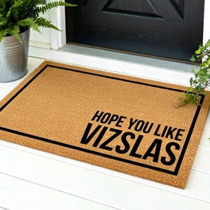 May include: A rectangular doormat with a brown surface and a black border. The text "HOPE YOU LIKE VIZSLAS" is printed in bold black letters. The mat is placed in front of a black door.