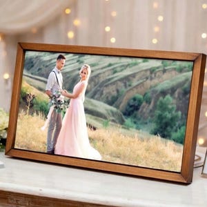 May include: A framed photograph of a couple in wedding attire, set against a scenic outdoor backdrop. The wooden frame has a warm, natural finish. The bride wears a pink dress, and the groom wears a light-colored shirt and suspenders. The photo is displayed on a white surface.