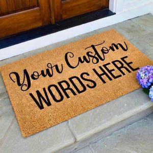May include: A rectangular doormat made of natural coir fiber, featuring the text "Your Custom WORDS HERE" in black script. The mat is placed on a stone step near a wooden door, with colorful flowers to the right.