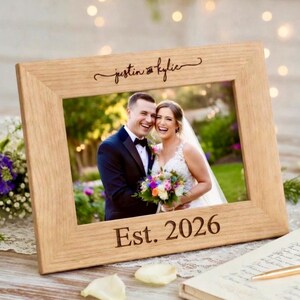 May include: A light-colored wooden picture frame with a wedding photo inside. The frame is engraved with the names "justin & kylie" and the date "Est. 2026". The photo shows a smiling couple, the bride holding a bouquet.