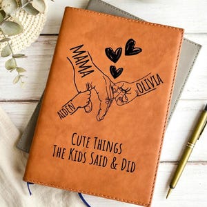May include: A tan leather journal with the words "MAMA", "AIDEN", and "OLIVIA" printed on it. The journal also has the text "Cute Things The Kids Said & Did" and a drawing of hands holding each other. A gold pen rests on the side.