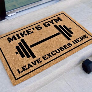 May include: A rectangular doormat with the text "MIKE'S GYM" above a barbell graphic and "LEAVE EXCUSES HERE" below. The mat is a natural brown color with black text and a black border. A black dumbbell is visible in the lower right corner.