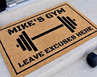 May include: A rectangular doormat with the text "MIKE'S GYM" above a barbell graphic and "LEAVE EXCUSES HERE" below. The mat is a natural brown color with black text and a black border. A black dumbbell is visible in the lower right corner.