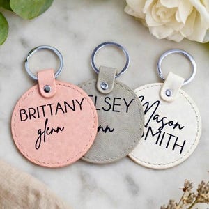 May include: Three personalized round keychains in pink, gray, and off-white. Each keychain features a name in black script. The keychains have silver metal rings and matching leather tabs. The background includes flowers and greenery.