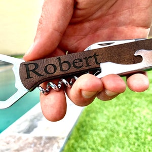 May include: A personalized wooden bottle opener and corkscrew with a silver bottle opener and corkscrew. The handle is engraved with the name "Robert".