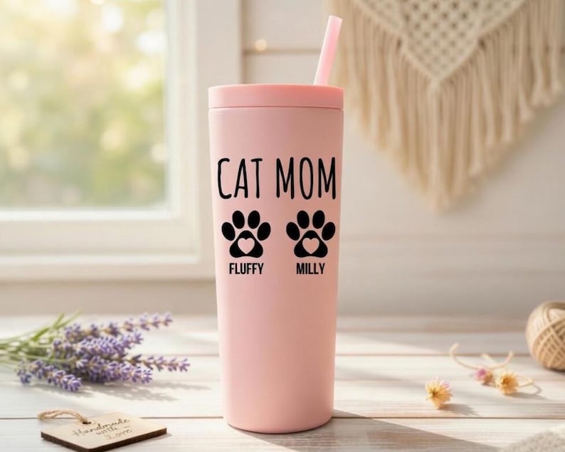 K&ouml;nnte beinhalten: Ein rosa Becher mit Deckel und Strohhalm. Auf dem Becher stehen die schwarzen Worte "CAT MOM" sowie zwei Pfotenabdr&uuml;cke mit Herzen. Unter den Pfotenabdr&uuml;cken stehen die Namen "Fluffy" und "Milly".