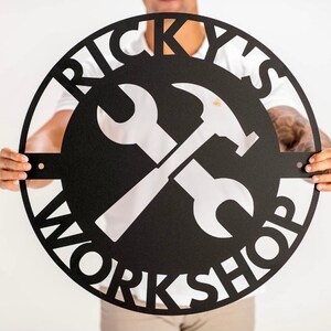 May include: Black metal sign with the words "Ricky's Workshop" in a circular design. A white wrench and hammer are crossed in the center.