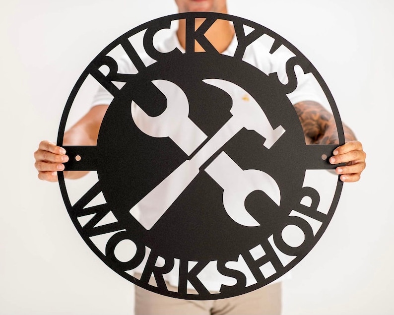 May include: Black metal sign with the words "Ricky's Workshop" in a circular design. A white wrench and hammer are crossed in the center.