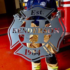 Op de afbeelding: Een zwart metalen brandweerembleem met de tekst "Est. 1984" en de naam "Henderson" in een gestileerd lettertype.