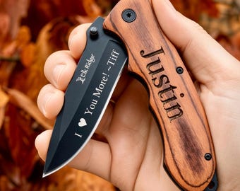 Puede incluir: Una navaja plegable con hoja negra y mango de madera veteada. La hoja presenta el texto "Elk Ridge" y "I ♥ You More! ~Tiff" en blanco. El mango de madera está grabado con el nombre "Justin" en una fuente oscura y negrita.