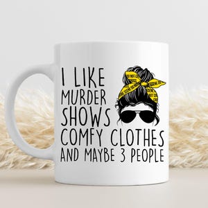 May include: White ceramic mug with a black and yellow design. The mug features a woman with a messy bun and a yellow ribbon that says "Do Not Cross Crime Scene Do Not Cross". The text on the mug reads "I like murder shows comfy clothes and maybe 3 people".