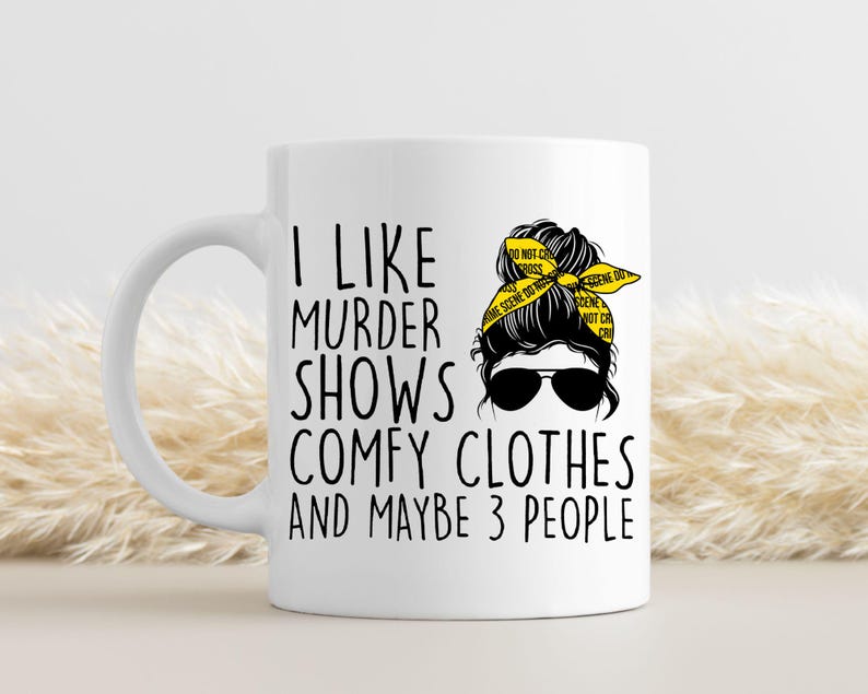 May include: White ceramic mug with a black and yellow design. The mug features a woman with a messy bun and a yellow ribbon that says "Do Not Cross Crime Scene Do Not Cross". The text on the mug reads "I like murder shows comfy clothes and maybe 3 people".