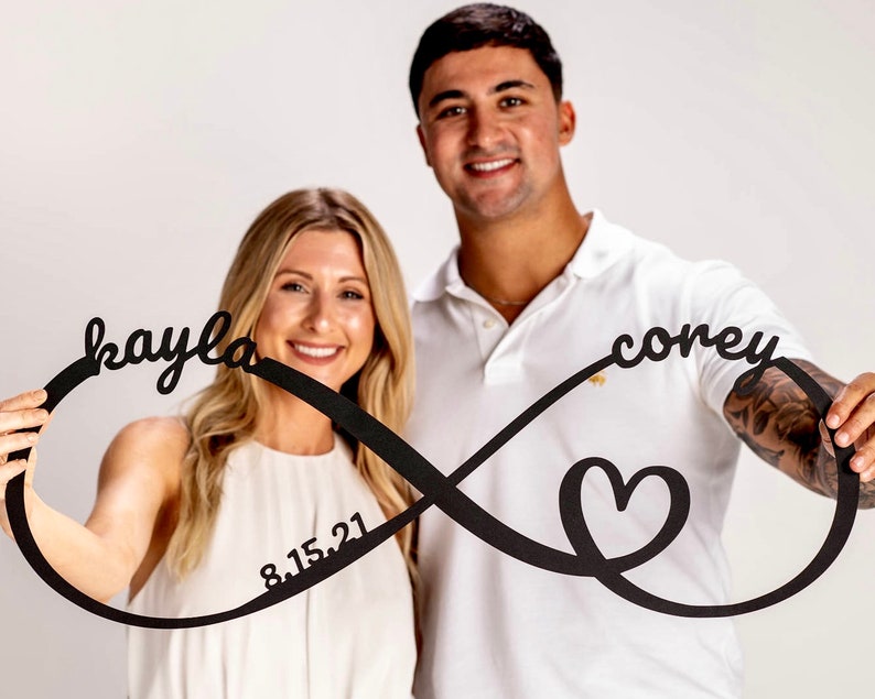 May include: A couple are holding a black infinity symbol with the names "kayla" and "corey" written on it. The symbol also includes a heart and the date "8.15.21".