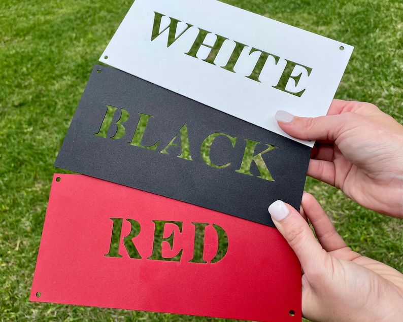 May include: Three metal signs with the words "WHITE", "BLACK", and "RED" cut out of the metal. The signs are stacked on top of each other, with the white sign on top, the black sign in the middle, and the red sign on the bottom.