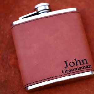 May include: A brown leather-covered flask with a silver metal top and bottom. The flask has the name "John" and the word "Groomsman" engraved on it.