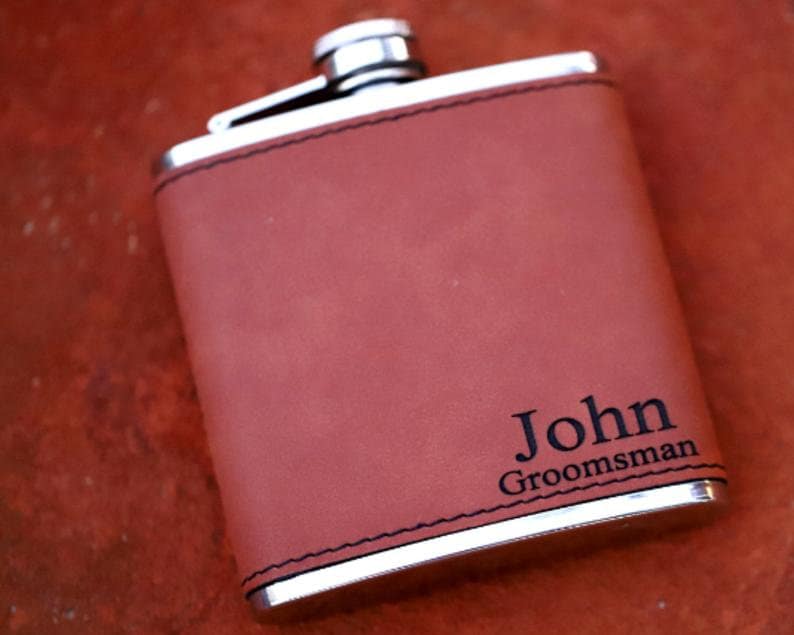 May include: A brown leather-covered flask with a silver metal top and bottom. The flask has the name "John" and the word "Groomsman" engraved on it.