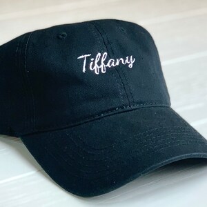 May include: Black baseball cap with a curved peak. The word "Tiffany" is embroidered in white script on the front. The cap is made of a durable fabric and has a classic, casual style.
