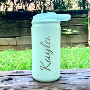 May include: A light green water bottle with a light blue lid and straw. The name "Kayla" is engraved on the side in a cursive font. The bottle is sitting on a wooden surface with a green background.