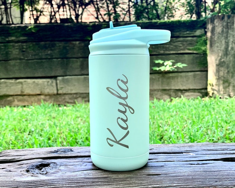 May include: A light green water bottle with a light blue lid and straw. The name "Kayla" is engraved on the side in a cursive font. The bottle is sitting on a wooden surface with a green background.