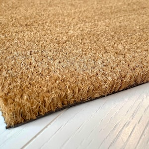 May include: Close-up of a rectangular doormat made of natural coir fibers. The mat has a light brown color and a textured surface. The bottom edge is lined with a black border. The doormat is placed on a white wooden surface.