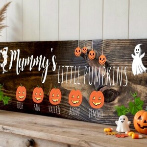 May include: A rustic wooden sign with the words "Mummy's Little Pumpkins" in white script. The sign features orange pumpkin illustrations and the names Tosh, Paul, Valerie, Brody, and Jimmy. A carved pumpkin and ghost decorations are also present.