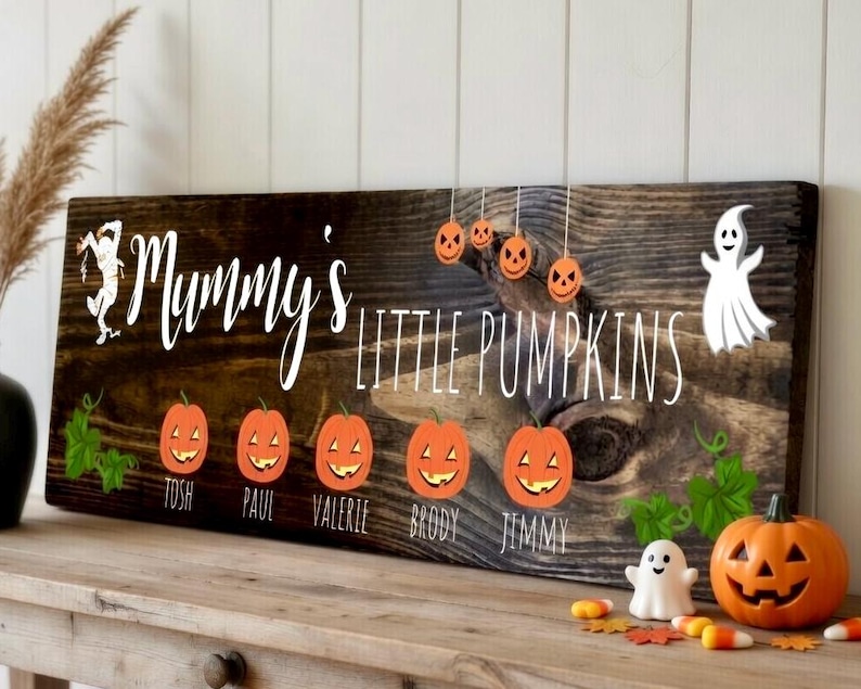 May include: A rustic wooden sign with the words "Mummy's Little Pumpkins" in white script. The sign features orange pumpkin illustrations and the names Tosh, Paul, Valerie, Brody, and Jimmy. A carved pumpkin and ghost decorations are also present.