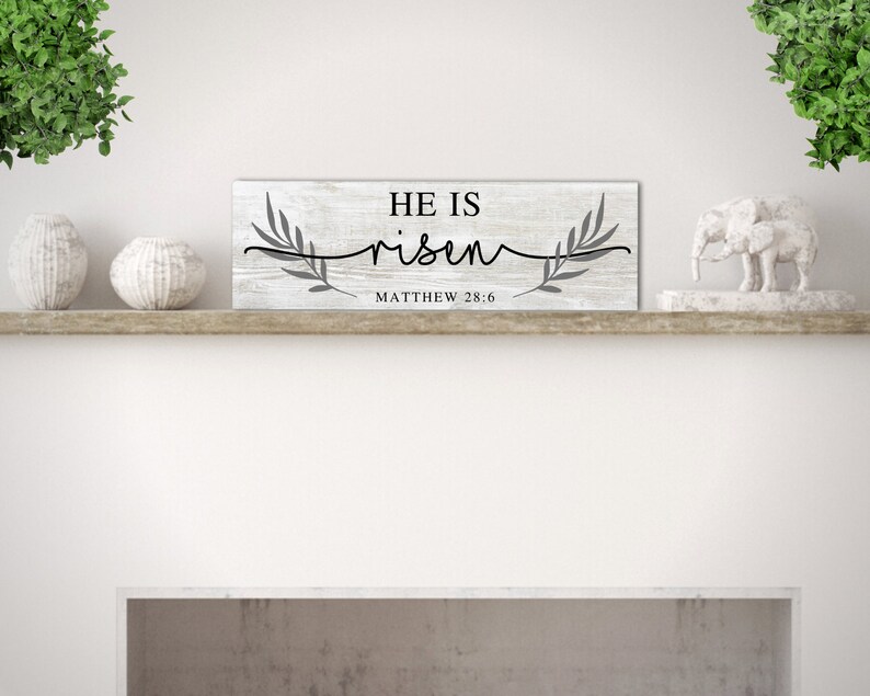 May include: White wooden sign with the text "He is risen" in black lettering and a gray floral design. The text "Matthew 28:6" is also included in black lettering.