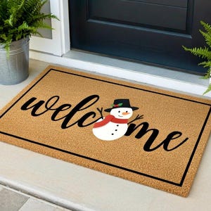 K&ouml;nnte beinhalten: Eine rechteckige Fu&szlig;matte mit einer braunen Kokosfaseroberfl&auml;che und einem schwarzen Rand. Das Wort "welcome" ist in schwarzen Schreibschriftbuchstaben geschrieben, mit einer Schneemann-Grafik. Der Schneemann hat einen schwarzen Hut, einen roten Schal und schwarze Stockarme.