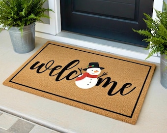 May include: A rectangular doormat with a brown coir surface and a black border. The word "welcome" is written in cursive black letters, with a snowman graphic. The snowman has a black hat, red scarf, and black stick arms.