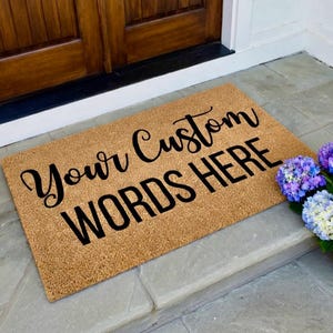 May include: A rectangular doormat made of natural coir fiber. The mat features the text "Your Custom WORDS HERE" in a black, cursive font. The mat is placed on a stone surface near a wooden door and a cluster of purple and blue hydrangeas.