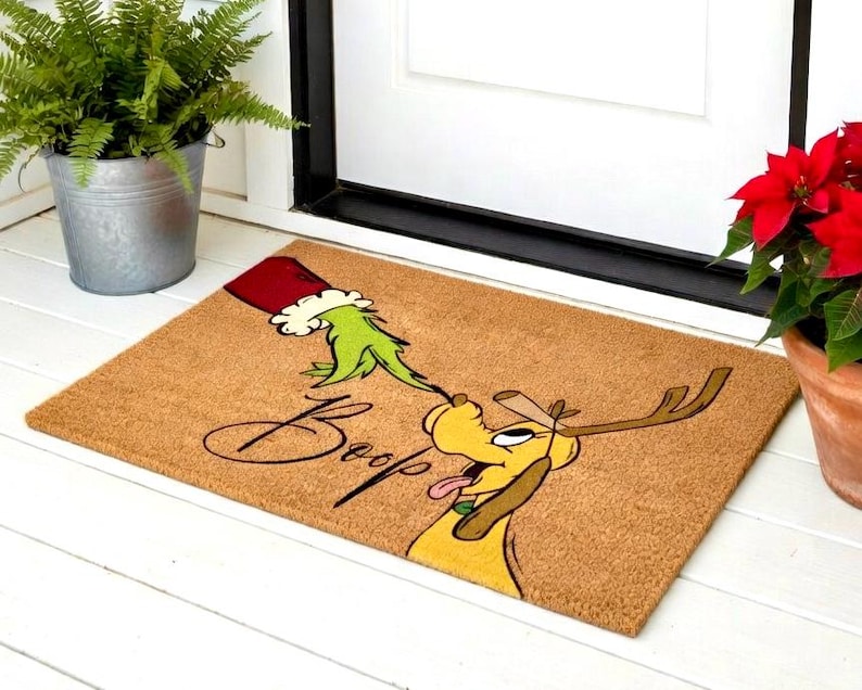 May include: A rectangular doormat with a brown background features a cartoon illustration. The design includes a green hand wearing a red and white Santa hat, reaching towards a yellow dog with antlers. The word "Boop" is written in cursive.