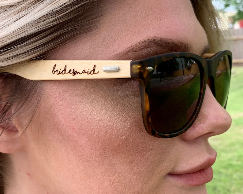 Pu&ograve; includere: Una persona indossa occhiali da sole marroni tartaruga con una montatura in legno che dice "bridesmaid" nella parte superiore.