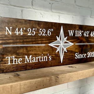 May include: A rectangular wooden sign with a dark brown stain. White text includes coordinates, a compass rose, the name "The Martin's", and the year "2026". The sign is displayed on a wooden shelf.