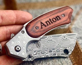May include: A silver pocket knife with a patterned blade and a wooden handle. The handle is engraved with the name "Anton".