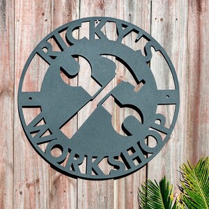 May include: A black metal sign with the words "Ricky's Workshop" in a circle with a wrench and hammer crossed in the center.