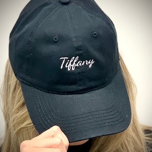 May include: A black baseball cap featuring the name "Tiffany" embroidered in white cursive script. The cap has a curved peak and an adjustable strap. The hat is made of a durable fabric, suitable for everyday wear.