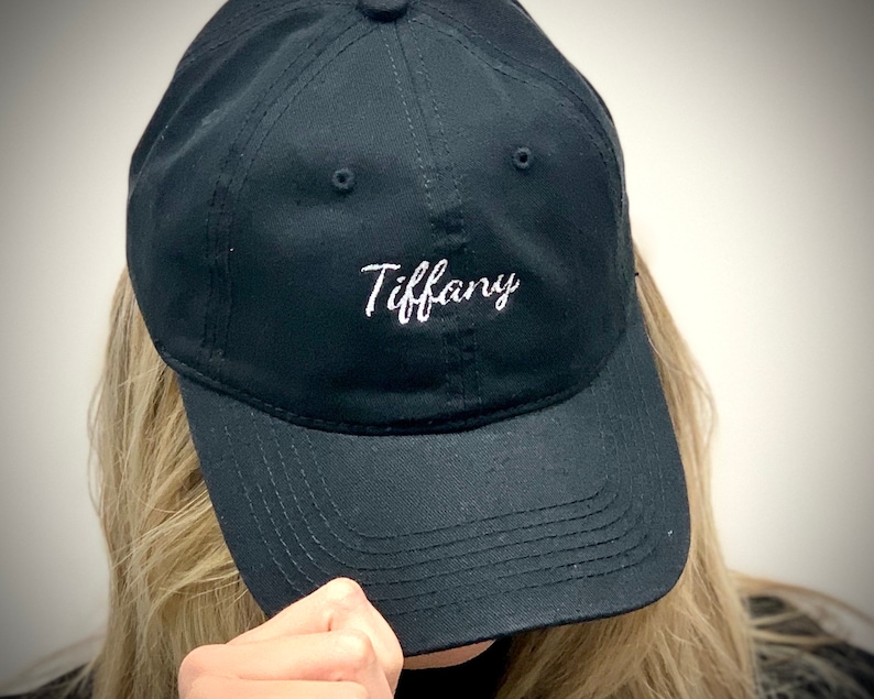 May include: A black baseball cap featuring the name "Tiffany" embroidered in white cursive script. The cap has a curved peak and an adjustable strap. The hat is made of a durable fabric, suitable for everyday wear.