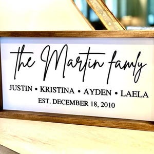 May include: A rectangular wooden sign with a white background and black text. The sign reads "the Martin family" in cursive, with the names Justin, Kristina, Ayden, and Laela below. The date "EST. DECEMBER 18, 2010" is also included.