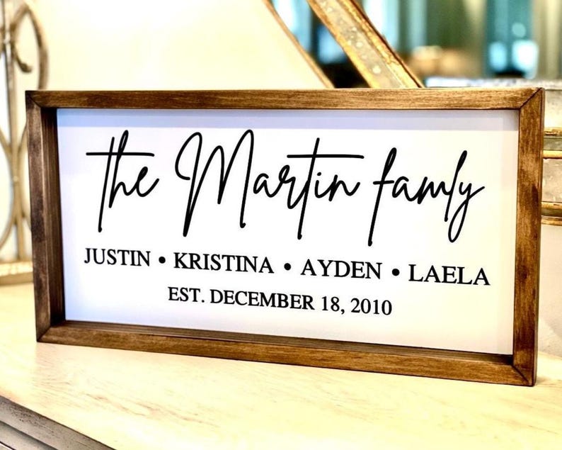 May include: A rectangular wooden sign with a white background and black text. The sign reads "the Martin family" in cursive, with the names Justin, Kristina, Ayden, and Laela below. The date "EST. DECEMBER 18, 2010" is also included.
