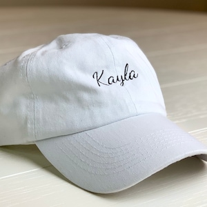 May include: A white baseball cap with a curved peak. The name "Kayla" is embroidered in black cursive on the front. The cap is made of a soft fabric and has a classic, casual style, suitable for everyday wear.