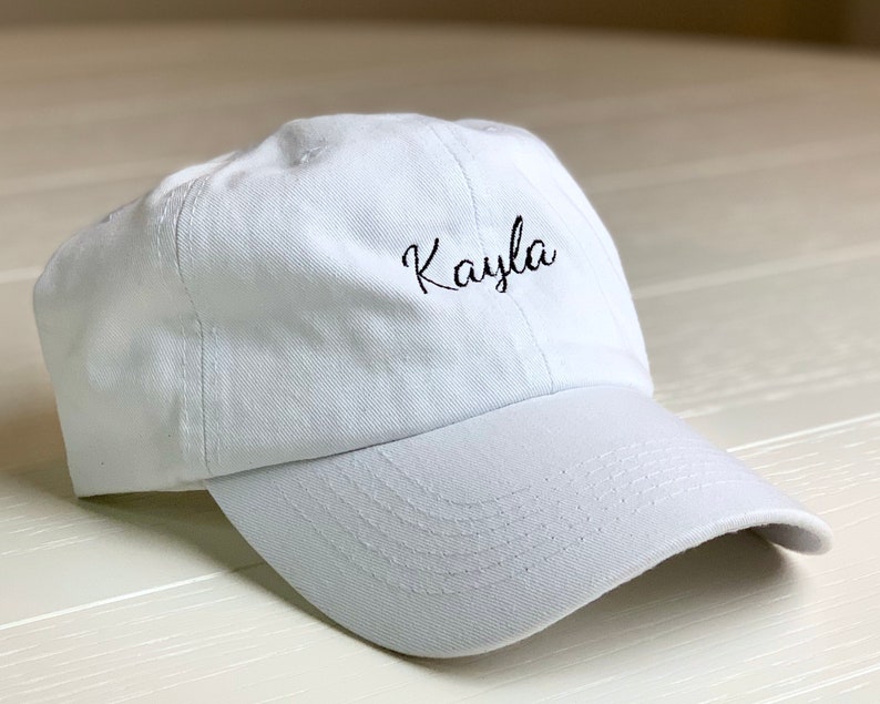 May include: A white baseball cap with a curved peak. The name "Kayla" is embroidered in black cursive on the front. The cap is made of a soft fabric and has a classic, casual style, suitable for everyday wear.