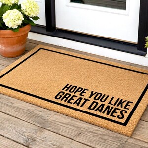 May include: A rectangular doormat made of natural coir fiber. The mat has a black border and the text "HOPE YOU LIKE GREAT DANES" in black, angled in the lower right corner. The mat is placed on a wooden porch.
