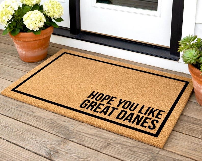 May include: A rectangular doormat made of natural coir fiber. The mat has a black border and the text "HOPE YOU LIKE GREAT DANES" in black, angled in the lower right corner. The mat is placed on a wooden porch.