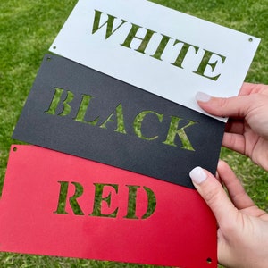 Puede incluir: Tres letreros de metal con las palabras "WHITE", "BLACK" y "RED" recortadas en el metal. Los letreros est&aacute;n apilados uno encima del otro, con el letrero blanco en la parte superior, el letrero negro en el medio y el letrero rojo en la parte inferior.