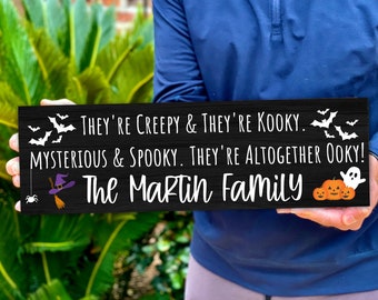 May include: Black Halloween sign with white text that reads "They're creepy & they're kooky. Mysterious & spooky. They're altogether ooky! The Martin Family". The sign is decorated with bats, a witch's hat, a spider, a ghost, and pumpkins.