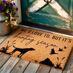 May include: A rectangular doormat with the text "WE'D LOVE TO, BUT IT'S hunting season" in black script. The mat features a black silhouette of a dog, birds, and foliage on a tan background. The mat is placed on a wooden porch.