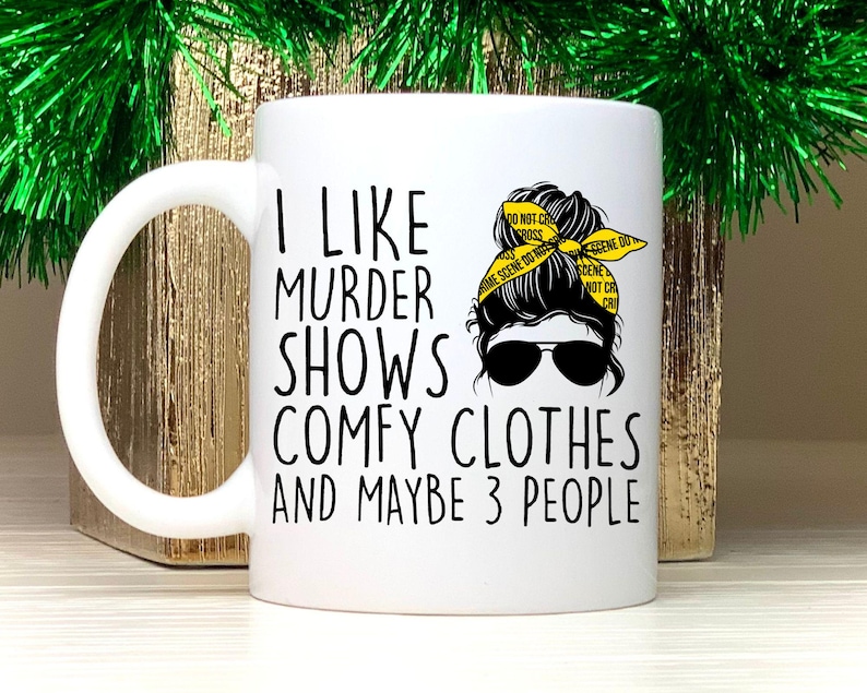 May include: White ceramic mug with a black and white graphic of a woman with a messy bun wearing a yellow "crime scene do not cross" headband. The text on the mug reads "I like murder shows comfy clothes and maybe 3 people."