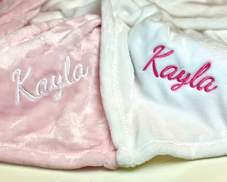 May include: Two personalized blankets, one pink with white embroidery and the other white with pink embroidery. Both blankets feature the name "Kayla" embroidered on them.