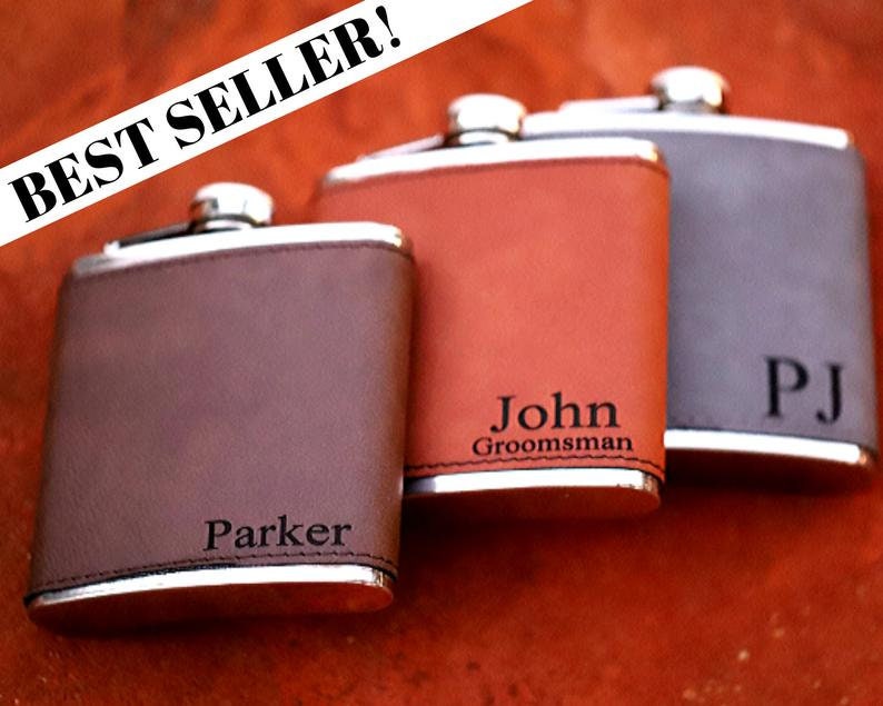 May include: Three personalized leather-wrapped stainless steel hip flasks. The flasks are brown, orange, and gray. The flasks are engraved with the names "Parker", "John Groomman", and "PJ".